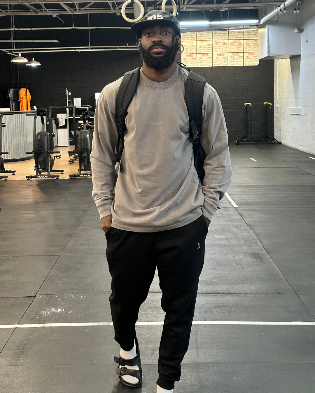 Man with a beard wearing a gray sweatshirt and black pants in a gym setting